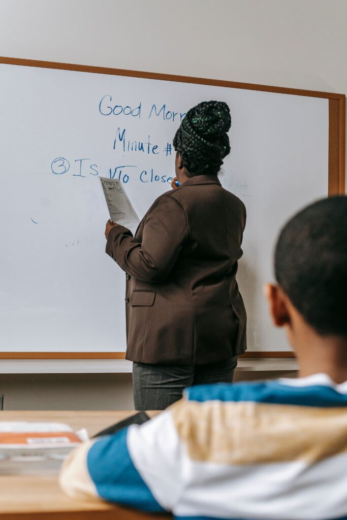 pexels-photo-5905467-5905467 Back view anonymous African American schoolboy sitting at desk in classroom and listening to female teacher taking notes on whiteboard